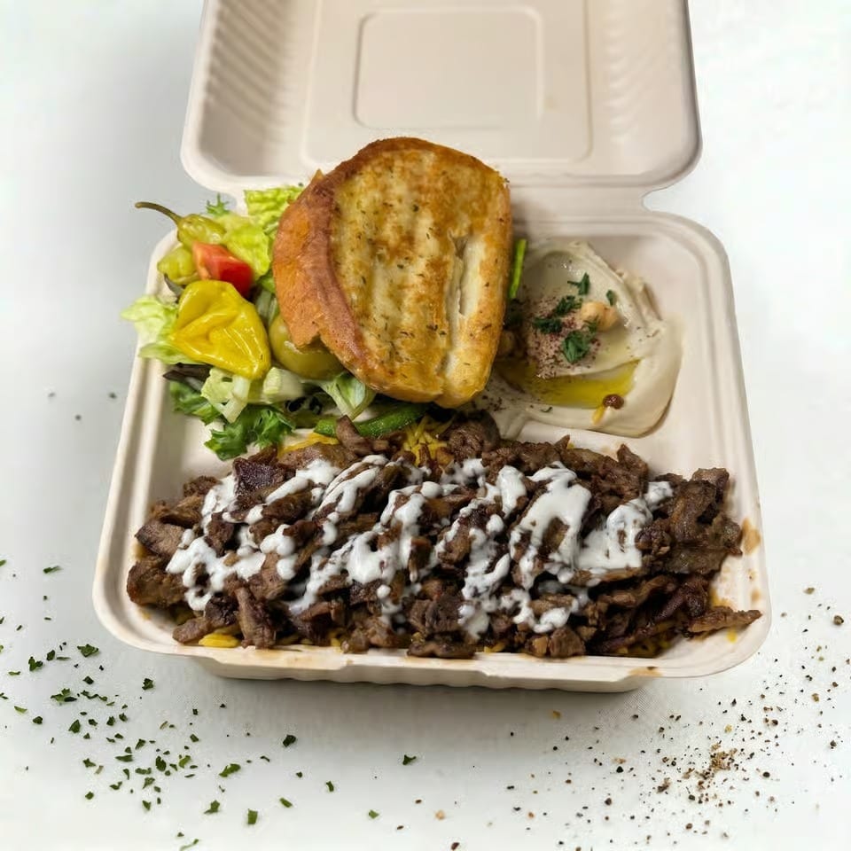 Takeout container with shawarma, hummus, salad, and toasted bread on a white surface.